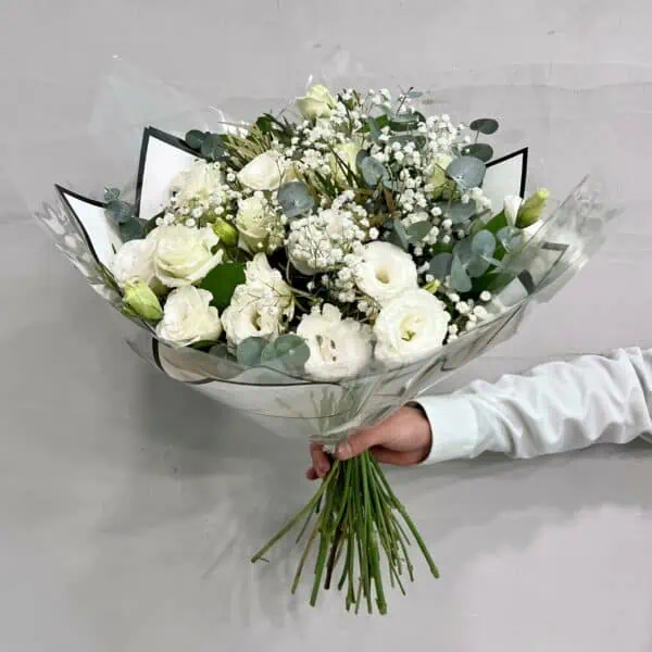 זר פרחים ליזי לבן הוא זר המבוסס על טהרת הצבע הלבן. שזור מפרחי ליזיאנטוס לבן, גיבסנית לבנה ואקליפטוס סילבר בשילוב ירק וענפי קישוט. הזר צולם מוחזק ביד על רקע הקיר בחנות.