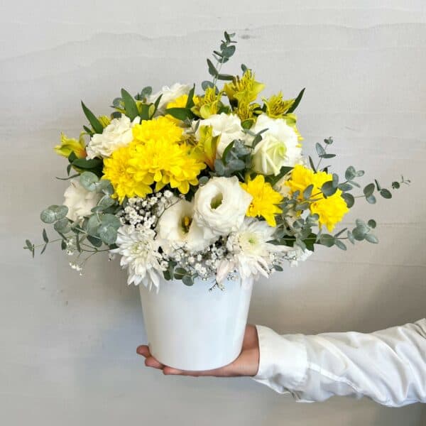 סידור פרחים מתנה מהשמש הו סידור פרחים בצבעים של צהוב ולבן. שזור מפרחי ליזיאנטוס לבן, חרציות צהובות ולבנות, גיבסנית לבנה, אלסטורמירה צהובה ואקליפטוס סילבר. צולם בתוך כלי לבן מוחזק ביד.
