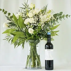 זר פרחים לבן שזור בתוך אגרטל שקוף לצד בקבוק יין מרלו. הזר שזור מפרחי שושן צחור, ליזיאנטוס לבן, גיבסנית לבנה בשילוב ירק וענפי קישוט.