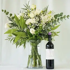 זר פרחים לבן שזור בתוך אגרטל שקוף לצד בקבוק יין שיראז. הזר שזור מפרחי שושן צחור, ליזיאנטוס לבן, גיבסנית לבנה בשילוב ירק וענפי קישוט.